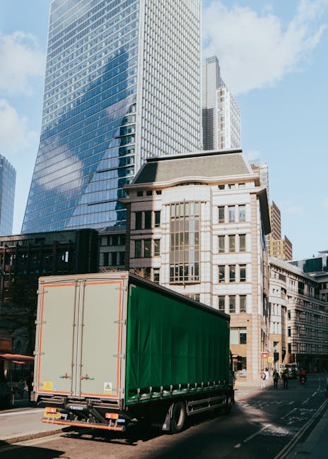 A large articulated truck with a green cargo container is parked on a city street during daytime, adjacent to modern high-rise office buildings with glass facades and older limestone structures. The truck's rear doors are closed, and it is positioned near a sidewalk where pedestrians and cyclists are visible. The surrounding environment includes construction or commercial activity, with the truck possibly involved in a home relocation or furniture transport process, as part of removal services in Kentish Town. The scene features natural daylight, with blue sky and scattered clouds overhead, highlighting the urban setting and the vehicle used for moving or delivery purposes, aligning with professional removals operations by Kentish Town Man and Van.