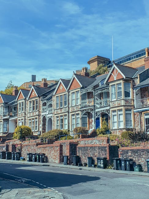 A row of Victorian-style terraced houses with dark brick facades, light-colored window frames, and pitched roofs lined along a quiet street under a clear blue sky. In the foreground, a well-maintained grassy area with a few leafless trees stretches across the image. The houses are set behind small front gardens enclosed by low fences. The scene is illuminated by bright daylight, highlighting the architectural details of the homes and the natural surroundings. The image relates to house removals and relocation services offered by Kentish Town Man and Van, which involves moving furniture and belongings between properties, often requiring careful planning, packing, and transportation logistics within residential areas.