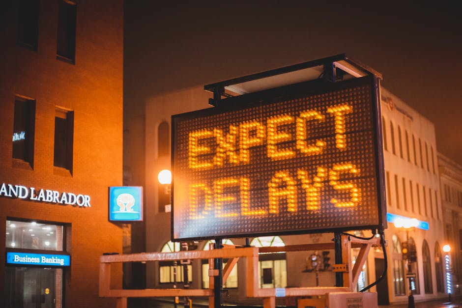 A large LED electronic signboard displaying the message 'EXPECT DELAYS' in bright orange lights is mounted on a metal frame and situated on a sidewalk near a commercial building at night. The background features partial views of a brick building with illuminated windows and signage, including a sign reading 'LAND LABRADOR' and a blue illuminated sign indicating 'Business Banking'. The area is lit by streetlights casting a warm glow, and the signboard is positioned close to the entryway of the nearby building, suggesting a busy urban environment. The scene reflects an alert related to transportation or service disruptions, consistent with logistics and home relocation contexts managed by companies like Kentish Town Man and Van, specializing in removals and moving services.
