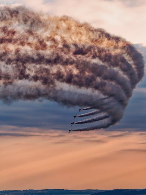 A formation of five aircraft flying in close proximity beneath an expansive, cloud-filled sky during sunset, leaving a trail of thick, dark smoke. The aircraft are aligned in a diagonal line from the lower left to the upper right, performing a precise manoeuvre often seen in air shows. The image captures the detailed smoke trails and the textured clouds, with a distant horizon at the bottom of the frame. This scene illustrates an impressive aerial display, similar to air show performances, which may be associated with special events or demonstrations involving skilled pilots and coordinated flying teams. The image is relevant to visual descriptions in the context of professional transportation or logistics, emphasizing the importance of precision and coordination, akin to home relocation or furniture transport processes handled by [COMPANY_NAME] as part of moving services.