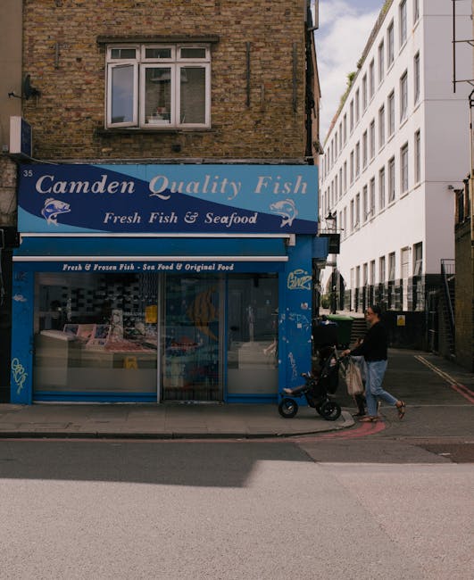 A street scene featuring a two-storey brick building with a blue signage board reading 'Camden Quality Fish' and 'Fresh Fish & Seafood' above a shop front with large glass windows showing seafood products inside. The shop appears to be related to fish and seafood sales. In front of the shop, a person dressed in casual clothing is pushing a stroller with a bag hanging from its handle, walking on the pavement. The area is well-lit with natural daylight, and nearby modern white apartment buildings are visible. The setting suggests an urban environment suitable for home relocation or moving services, with the image capturing the process of loading or transporting items, typical of furniture transport or packing and moving activities often managed by companies like Kentish Town Man and Van.