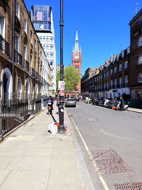 A wide street view in Kentish Town showing a row of traditional brick terraced houses with white window frames and small front gardens on the right side, and modern multi-storey buildings with reflective glass facades on the left side. A black streetlamp is positioned in the foreground, with a sidewalk along the street where a person is walking next to a pile of white cardboard boxes and other packing materials. Several parked scooters and cars are lined up along the curb, and a few pedestrians and vehicles are visible in the distance. The sky is clear and blue, with a prominent red clock tower of a historic building, possibly a church, visible at the end of the street. This scene depicts a typical urban environment suitable for house removals and furniture transport, as conducted by Kentish Town Man and Van, with ongoing loading and moving logistics evident in the street scene.