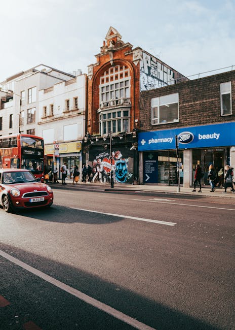 A busy urban street scene in Kentish Town with a row of commercial buildings including a pharmacy and beauty store on the ground floor, featuring large shopfront windows and signage. In front, pedestrians are walking on the sidewalk, some crossing the street, while others stand near storefronts. A red double-decker bus is seen stopping at a bus stop on the left side, and a red car drives along the road in the foreground. The buildings above consist of a mix of architectural styles, with a prominent brick structure at the center that has a large arched window and a decorative tower-like feature at the top. The street appears to be part of a home relocation or moving logistics scene, and it is captured in daylight under a partly cloudy sky, with ambient lighting illuminating the facade and street activity. Kentish Town Man and Van, a removal service, would be familiar with the busy street environment suitable for furniture transport and packing and moving operations, particularly in relation to Camden Council permit rules for moving vans.