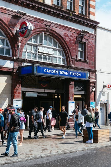 A busy urban street scene in Kentish Town with a row of commercial buildings including a pharmacy and beauty store on the ground floor, featuring large shopfront windows and signage. In front, pedestrians are walking on the sidewalk, some crossing the street, while others stand near storefronts. A red double-decker bus is seen stopping at a bus stop on the left side, and a red car drives along the road in the foreground. The buildings above consist of a mix of architectural styles, with a prominent brick structure at the center that has a large arched window and a decorative tower-like feature at the top. The street appears to be part of a home relocation or moving logistics scene, and it is captured in daylight under a partly cloudy sky, with ambient lighting illuminating the facade and street activity. Kentish Town Man and Van, a removal service, would be familiar with the busy street environment suitable for furniture transport and packing and moving operations, particularly in relation to Camden Council permit rules for moving vans.