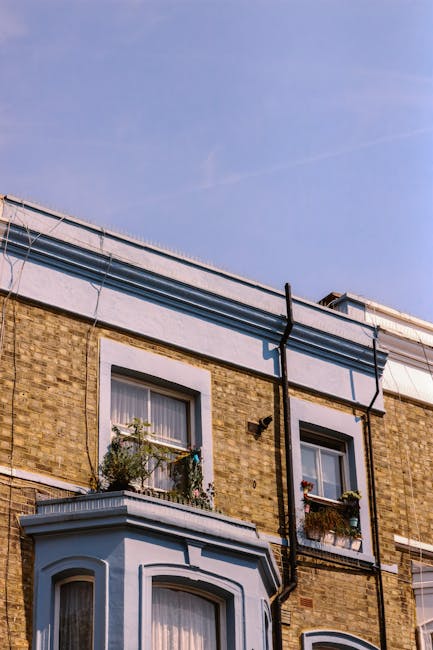 The image shows the exterior of a multi-storey brick building with two visible windows on the second floor, each adorned with flower boxes containing small plants and flowers. The building's facade features a decorative white trim around the windows and a curved bay window on the ground floor. A black downpipe runs vertically along the wall, connecting to the guttering system at the roofline. The roof's edge is visible, with part of the white fascia and guttering in view. The sky above is clear with a slight blue hue, indicating daylight. This setting depicts a residential property characteristic of London housing, potentially viewed during a home relocation or moving process. Kentish Town Man and Van provides professional removals services that may involve navigating such exterior features during furniture transport or packing and moving tasks, especially when planning safe access for loading or unloading items from elevated windows or entrances.