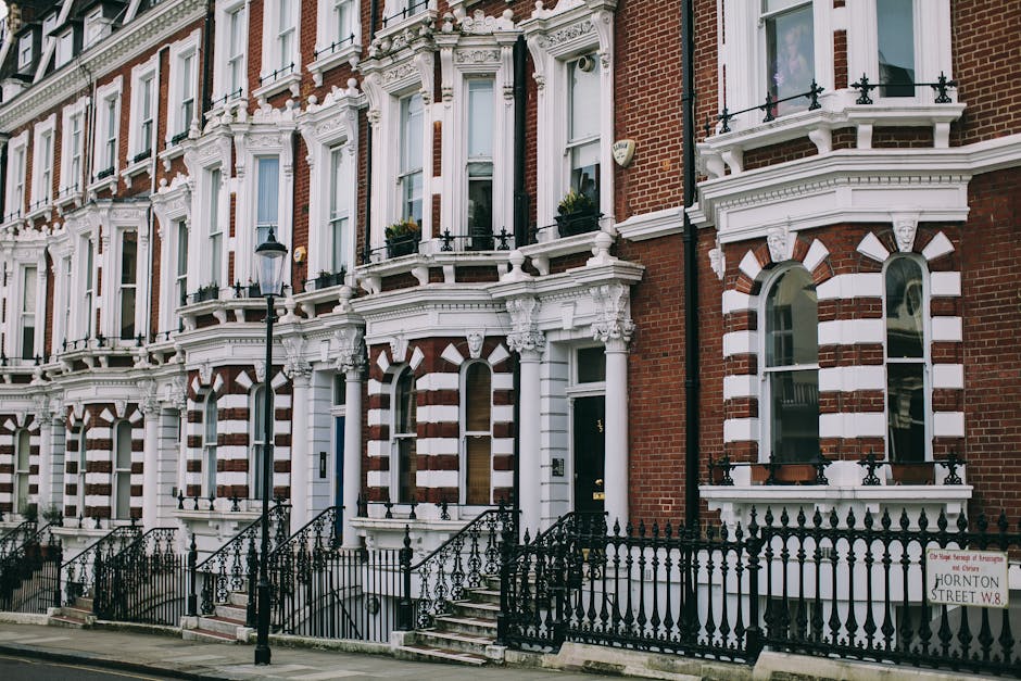 A row of Victorian-style terraced houses with red brick facades and white decorative architectural details, including ornate window frames, cornices, and balustrades. The buildings feature large, arched ground-floor windows with white trim and smaller, rectangular upper windows, some with window boxes. In front of the houses, black wrought iron fences and stair railings lead to small steps up to the entrances. A street lamp stands near the pavement, and a sign indicates the address as Hornton Street W8. The scene captures the exterior of residential properties, providing a suitable visual context for house removals and home relocation services, with potential footage of moving or packing activities occurring inside or in front of these homes, as indicated by the context of Kentish Town house removals.