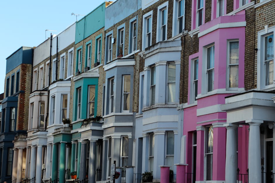A row of Victorian terraced houses in Kentish Town with colourful facades painted in shades of blue, white, and pink. The buildings feature bay windows, some with flower boxes, and wrought iron railings along the pavement. The ground floors show doorways and small front gardens, with a partially visible van parked nearby, suggesting a relocation or house removal process. Exterior lighting and a clear sky indicate daytime. This setting illustrates typical residential architecture suitable for house removals and furniture transport, as handled by Kentish Town Man and Van, during packing, loading, and home relocation activities.