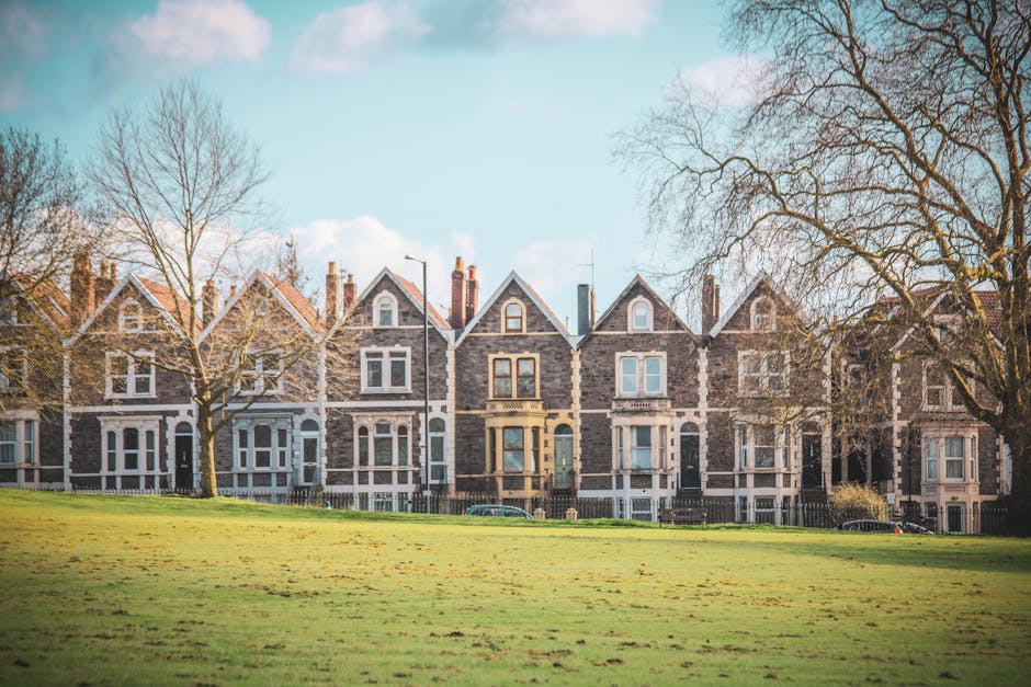 A row of Victorian terraced houses with grey stone facades, white window frames, and pitched roofs, located overlooking a large grassy park area. Several leafless trees are situated near the houses, with some branches extending over the lawn. The sky is partly cloudy with patches of blue visible. In front of the houses, a pavement runs parallel to the street where two cars are parked. The scene suggests a peaceful residential neighbourhood. This image represents typical London Victorian terraces, relevant to Kentish Town home removals and house relocation services provided by Kentish Town Man and Van, including furniture transport and packing during a residential moving process.