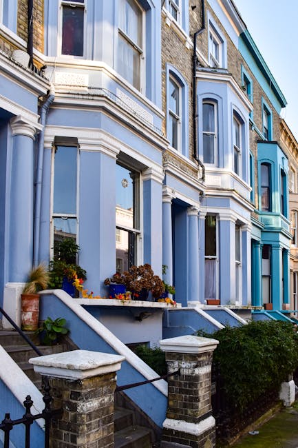 A row of Victorian terraced houses in Kentish Town with colourful facades painted in shades of blue, white, and pink. The buildings feature bay windows, some with flower boxes, and wrought iron railings along the pavement. The ground floors show doorways and small front gardens, with a partially visible van parked nearby, suggesting a relocation or house removal process. Exterior lighting and a clear sky indicate daytime. This setting illustrates typical residential architecture suitable for house removals and furniture transport, as handled by Kentish Town Man and Van, during packing, loading, and home relocation activities.