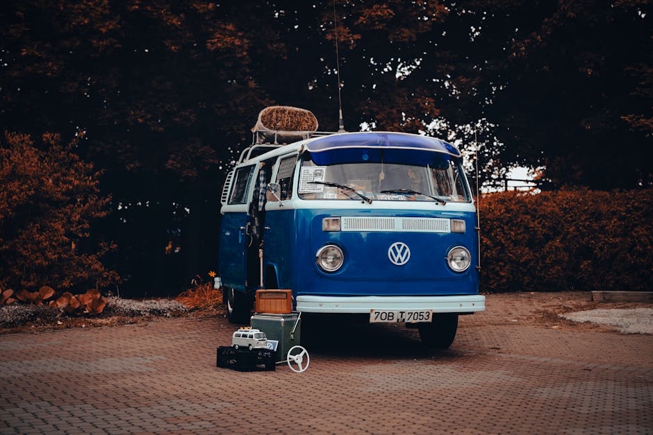 A vintage blue Volkswagen Transporter van parked on a paved area with a license plate reading '70B T 7053', surrounded by a few small suitcases, a black camera, and a white steering wheel toy. The van's roof carries a large, rolled-up fabric item secured with a strap, and its windows reflect the dark trees and sky behind it. The vehicle is positioned outside, likely prepared for a home relocation or furniture transport, with the background featuring leafy trees and bushes. The scene depicts the loading or transit stage of a moving process, associated with house removals and packing services, as offered by Kentish Town Man and Van. The environment appears calm, with soft lighting suggesting early morning or late afternoon, emphasizing the focus on the vehicle and packed items in the context of moving logistics.