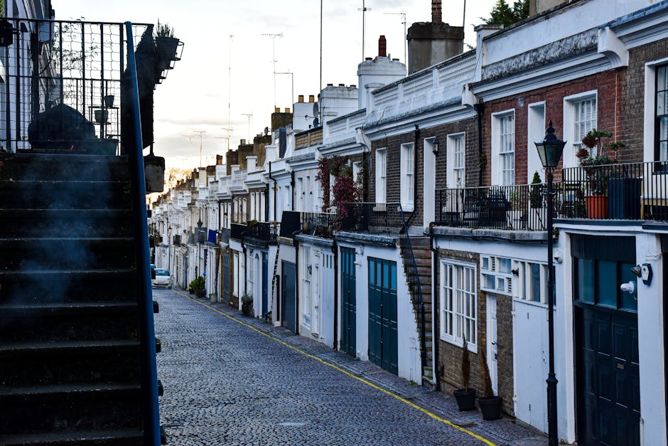 A narrow residential street in London with a line of terraced houses featuring brick and white-painted facades, some with small front gardens adorned with potted plants and decorative lanterns. The houses have traditional sash windows, with one property showing a black external staircase leading up to the doorway. The street is cobbled, with a distinct yellow line marking the edge of the pavement. On the left side, there is a black metal staircase, possibly leading to an upper-level flat, with a plastic bag placed on the steps. The scene is taken during the late afternoon or early evening, with soft natural light illuminating the buildings. This setting illustrates a typical environment for home relocation or furniture transport services, as it highlights narrow passageways and staircases often encountered in house removals, supported by companies like Kentish Town Man and Van for packing, loading, and transportation tasks.