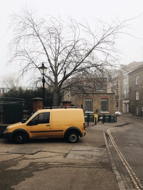 A vintage blue Volkswagen Transporter van parked on a paved area with a license plate reading '70B T 7053', surrounded by a few small suitcases, a black camera, and a white steering wheel toy. The van's roof carries a large, rolled-up fabric item secured with a strap, and its windows reflect the dark trees and sky behind it. The vehicle is positioned outside, likely prepared for a home relocation or furniture transport, with the background featuring leafy trees and bushes. The scene depicts the loading or transit stage of a moving process, associated with house removals and packing services, as offered by Kentish Town Man and Van. The environment appears calm, with soft lighting suggesting early morning or late afternoon, emphasizing the focus on the vehicle and packed items in the context of moving logistics.