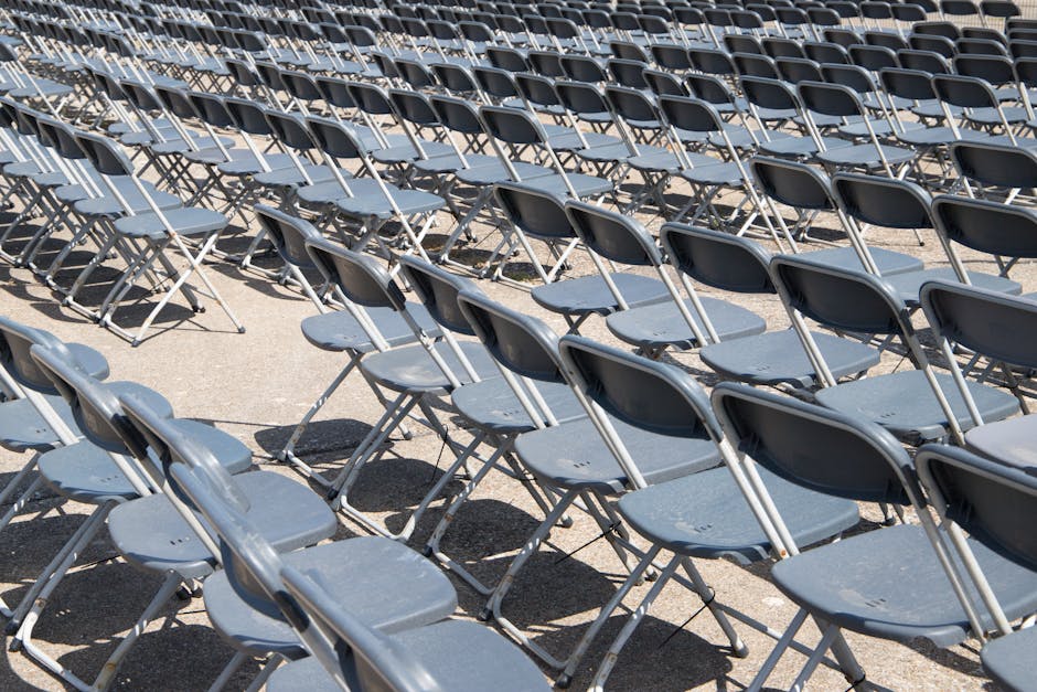 A large outdoor area filled with numerous black folding chairs arranged in rows on a paved surface, with some chairs slightly tilted or overlapping. The chairs are made of metal frames with plastic seats and backs, positioned under bright sunlight which casts distinct shadows on the ground. The setting appears to be prepared for an event, possibly in a public space or outdoor venue. This scene illustrates the types of equipment involved in event furniture setup or removal, relevant to home relocation and furniture transport services offered by Kentish Town Man and Van, particularly when managing the logistics of moving or storing event equipment as part of a broader house removal or rearrangement project.