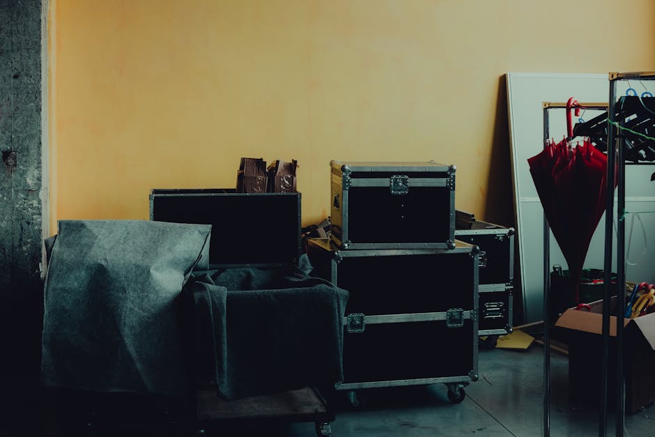 Interior scene showing a cluttered room with various packed items ready for home relocation, including black flight cases stacked on top of each other, a large wrapped piece of furniture covered in dark fabric or padding, and an open cardboard box filled with assorted tools and packing materials. A white wardrobe or storage unit is positioned against a yellow wall, with several red umbrellas hanging from a rack nearby. The room's floor appears to be concrete, and a partially visible door frame is on the left side of the image. The overall setting suggests an ongoing furniture transport or packing process, with the items prepared for loading into a van for removals services, as provided by Kentish Town Man and Van.