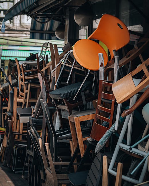A large outdoor area filled with numerous black folding chairs arranged in rows on a paved surface, with some chairs slightly tilted or overlapping. The chairs are made of metal frames with plastic seats and backs, positioned under bright sunlight which casts distinct shadows on the ground. The setting appears to be prepared for an event, possibly in a public space or outdoor venue. This scene illustrates the types of equipment involved in event furniture setup or removal, relevant to home relocation and furniture transport services offered by Kentish Town Man and Van, particularly when managing the logistics of moving or storing event equipment as part of a broader house removal or rearrangement project.