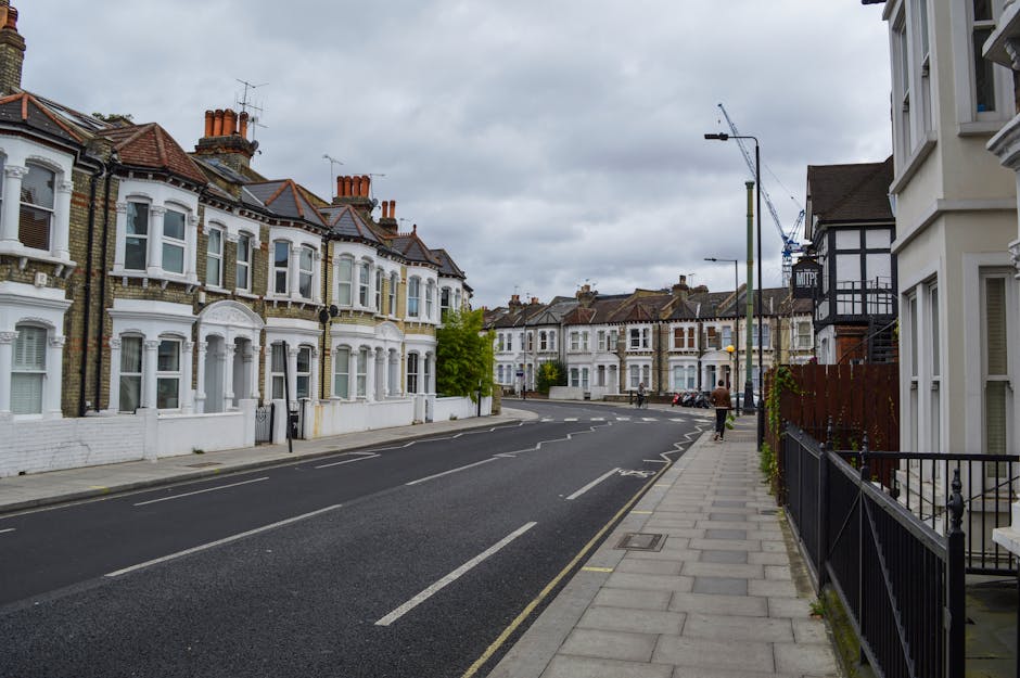 A quiet residential street in Kentish Town lined with rows of Victorian-style terraced houses, featuring white facades, bay windows, and tiled roofs under an overcast sky. The pavement runs alongside