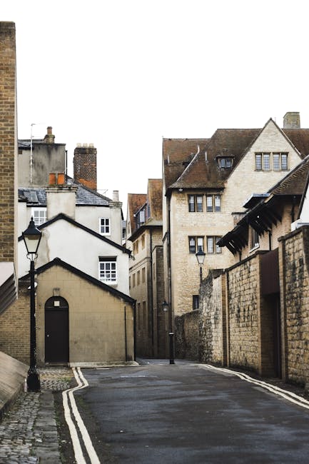 A narrow cobbled street or alleyway in a residential area during daytime, flanked by old multi-storey buildings with varied architectural styles, including brick and stone facades, small windows, and pitched roofs. Two black street lamps are mounted on the walls, and a small, detached single-storey structure with a pitched roof is situated on the left side. The street appears to be the setting for a house removal or home relocation, with signage for Kentish Town Man and Van, indicating a professional removal service. The scene is empty, with no vehicles or people visible, suggesting the area is prepared for ongoing furniture transport or packing and moving tasks related to house removals, typical of services offered in the Kentish Town area. The overcast sky provides diffuse natural light, highlighting the textured surfaces of the buildings and the street surface.