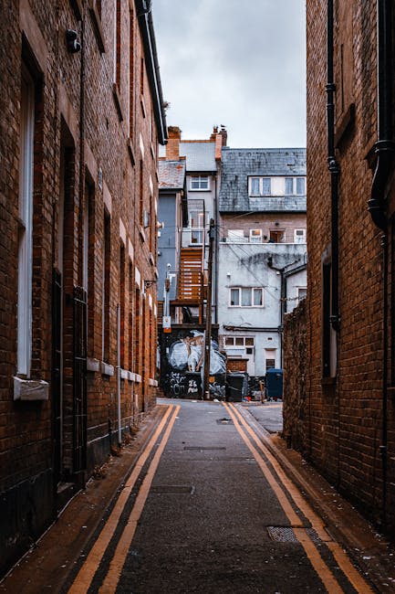 A narrow alleyway between two brick buildings with fire escapes and small windows. The ground is paved with asphalt, featuring double yellow lines along the edges. Towards the back of the alley, there are covered objects and waste bins, indicating a service or back entrance area. The scene is lit by natural daylight, with a cloudy sky visible above. This setting is typical for urban house removals or furniture transport in tight city spaces, where careful navigation is required. Kentish Town Man and Van specializes in such home relocation logistics, including loading and unloading processes through limited access points, ensuring efficient moving services in areas like Kentish Town.