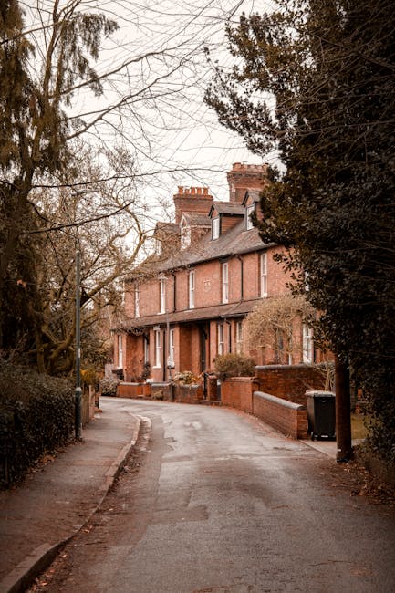 A narrow cobbled street or alleyway in a residential area during daytime, flanked by old multi-storey buildings with varied architectural styles, including brick and stone facades, small windows, and pitched roofs. Two black street lamps are mounted on the walls, and a small, detached single-storey structure with a pitched roof is situated on the left side. The street appears to be the setting for a house removal or home relocation, with signage for Kentish Town Man and Van, indicating a professional removal service. The scene is empty, with no vehicles or people visible, suggesting the area is prepared for ongoing furniture transport or packing and moving tasks related to house removals, typical of services offered in the Kentish Town area. The overcast sky provides diffuse natural light, highlighting the textured surfaces of the buildings and the street surface.