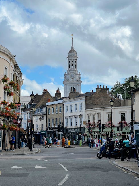 A busy town square in Kentish Town with a mix of commercial and residential buildings, including shops with awnings and flower baskets hanging outside. In the background, there is a tall, historic white clock tower with a spire topped by a weather vane, set against a partly cloudy sky. The street is populated with pedestrians walking on the pavements, some standing near the shop entrances, and others crossing the road. Several vehicles, including a black scooter and parked cars, are visible along the side of the street. The scene captures the typical environment of an active urban area where house removals and furniture transport services, such as those offered by Kentish Town Man and Van, operate around local relocation projects. The street lighting and signage indicate a well-established community, with some greenery seen in the form of potted plants and hanging flowers enhancing the local atmosphere.