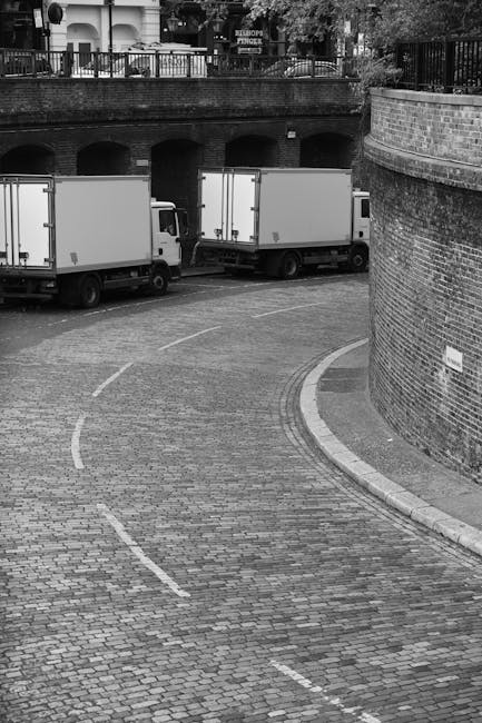 Two white box trucks are parked on a cobblestone driveway near a brick wall and an arched underpass, with residential buildings and trees visible in the background. The trucks are positioned side by side, with the rear doors of both vehicles closed. The driveway curves to the right and is marked with dashed white lines for parking or loading zones. The area appears to be an outdoor setting, likely within an urban or residential environment suitable for home relocations or furniture transport. Kentish Town Man and Van's removals service may utilize such vehicles for home relocation or moving logistics, including loading and unloading household items, furniture, and packing materials during a scheduled move in Kentish Town.