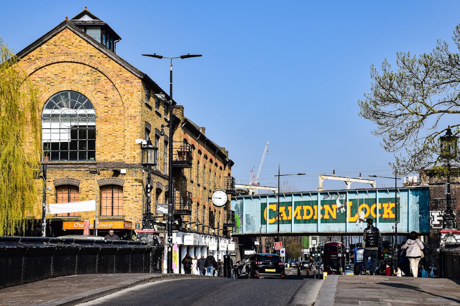 A busy town square in Kentish Town with a mix of commercial and residential buildings, including shops with awnings and flower baskets hanging outside. In the background, there is a tall, historic white clock tower with a spire topped by a weather vane, set against a partly cloudy sky. The street is populated with pedestrians walking on the pavements, some standing near the shop entrances, and others crossing the road. Several vehicles, including a black scooter and parked cars, are visible along the side of the street. The scene captures the typical environment of an active urban area where house removals and furniture transport services, such as those offered by Kentish Town Man and Van, operate around local relocation projects. The street lighting and signage indicate a well-established community, with some greenery seen in the form of potted plants and hanging flowers enhancing the local atmosphere.