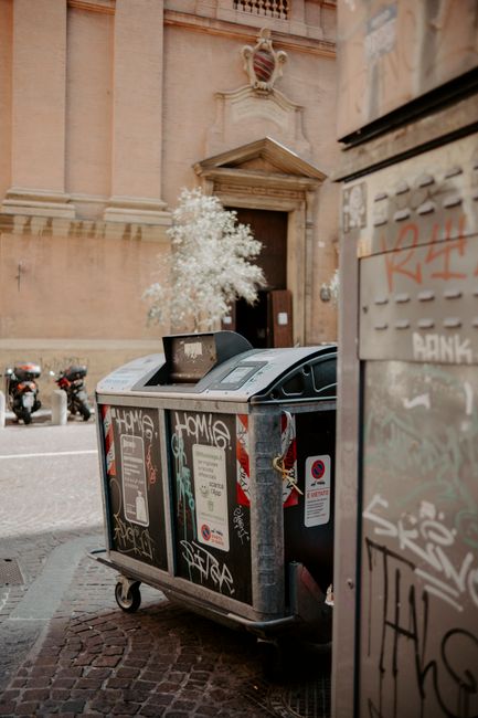 A large, grey, metal waste disposal container with graffiti markings on its surface is positioned on a cobbled pavement in front of a historic building with a stone doorway and decorative crest above